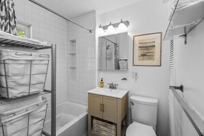 Une salle de bain avec une baignoire, des toilettes et un lavabo avec un miroir au-dessus. On y trouve également un porte-serviettes et un tableau encadré accroché au mur.