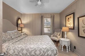 Une chambre à coucher avec un grand lit, une chaise, une table de chevet, un ventilateur de plafond et une fenêtre avec des stores.