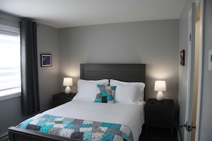 A neatly made bed with a quilt, two bedside tables with lamps, a window with curtains, and a door.
