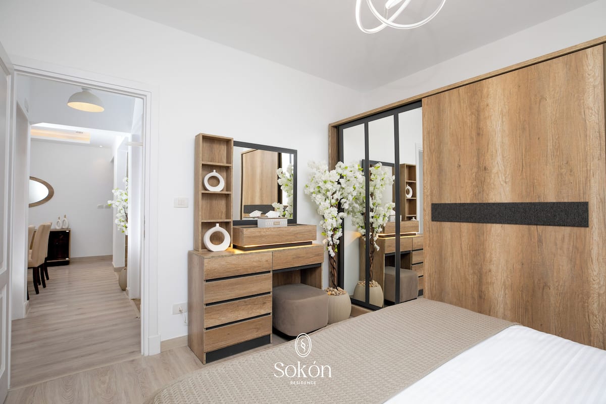 A modern bedroom with a wooden wardrobe, a vanity with a mirror, and a bedside table.