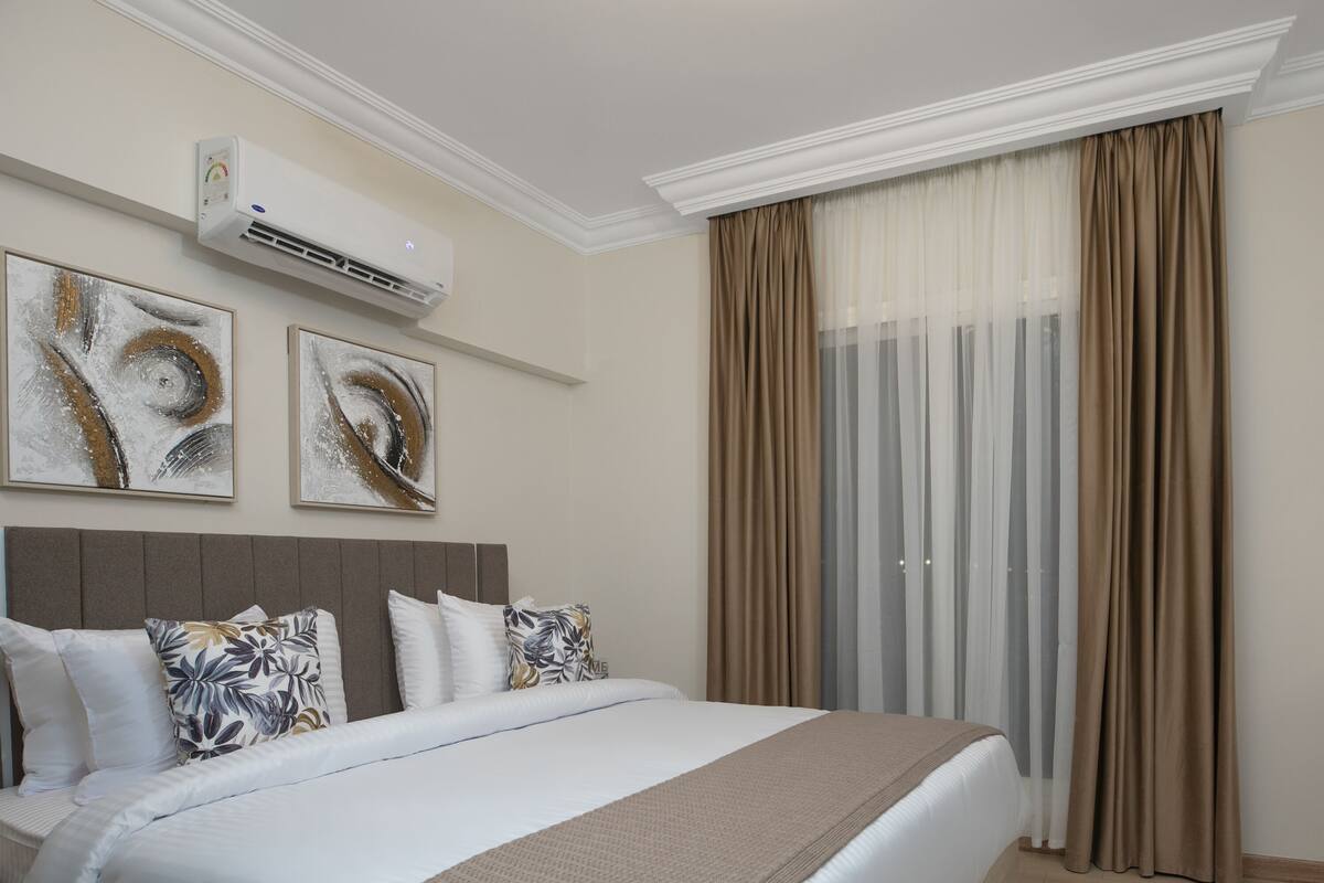 A hotel room with a bed, air conditioning unit, artwork, curtains, a television, and a small table with a vase.