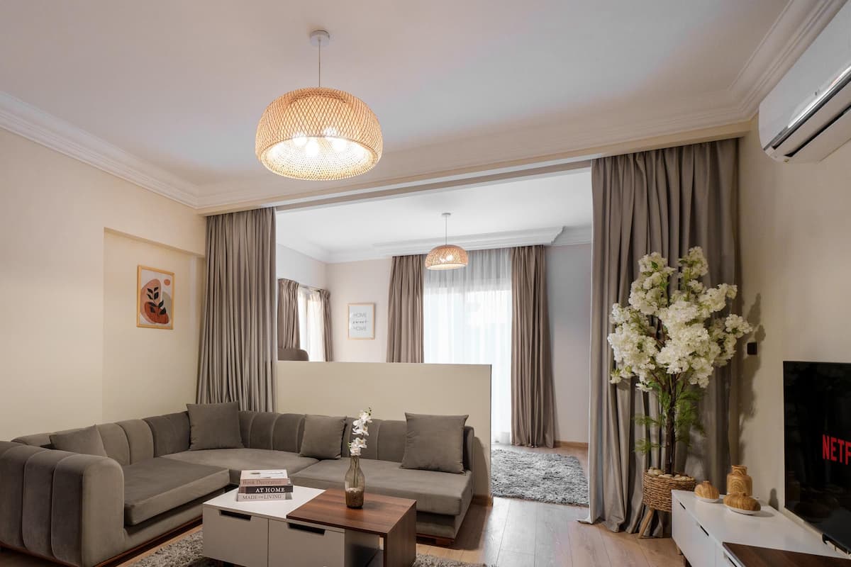 A modern living room with a sectional sofa, a coffee table, and a TV. There is a large mirror on the wall and a window with curtains.