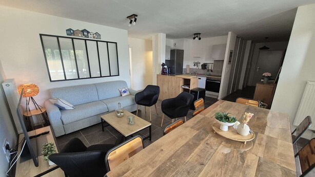A modern living room with a sofa, coffee table, and dining table. There is a kitchen in the background with a stove and cabinets.
