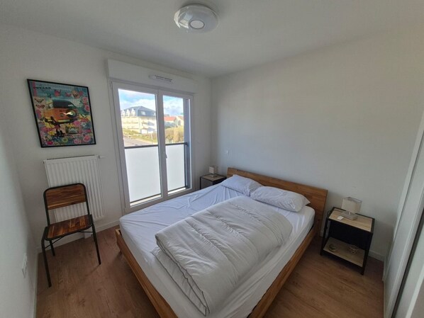 A bedroom with a bed, a chair, a nightstand, a radiator, and a window with a view of buildings. - 3-bedroom apartment in Stella-plage with parking and pets allowed (Cucq-Stella Plage)