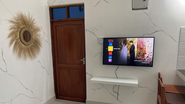 Una habitación con puerta de madera, un televisor de pared mostrando una película y un espejo decorativo con diseño de sol.