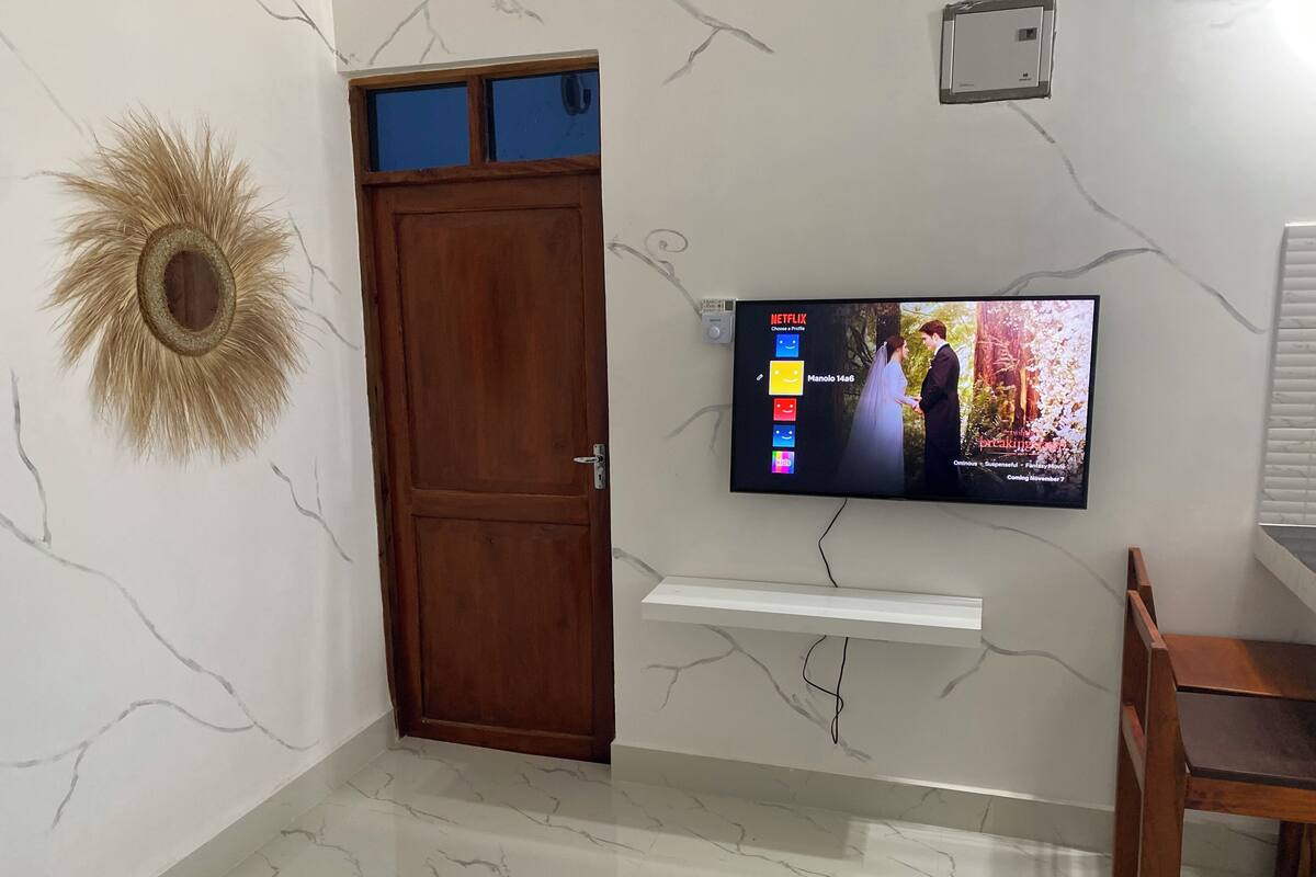 Ein Zimmer mit Holztür, einem an der Wand befestigten Fernseher, der einen Film zeigt, und einem dekorativen Spiegel in Sonnenform.