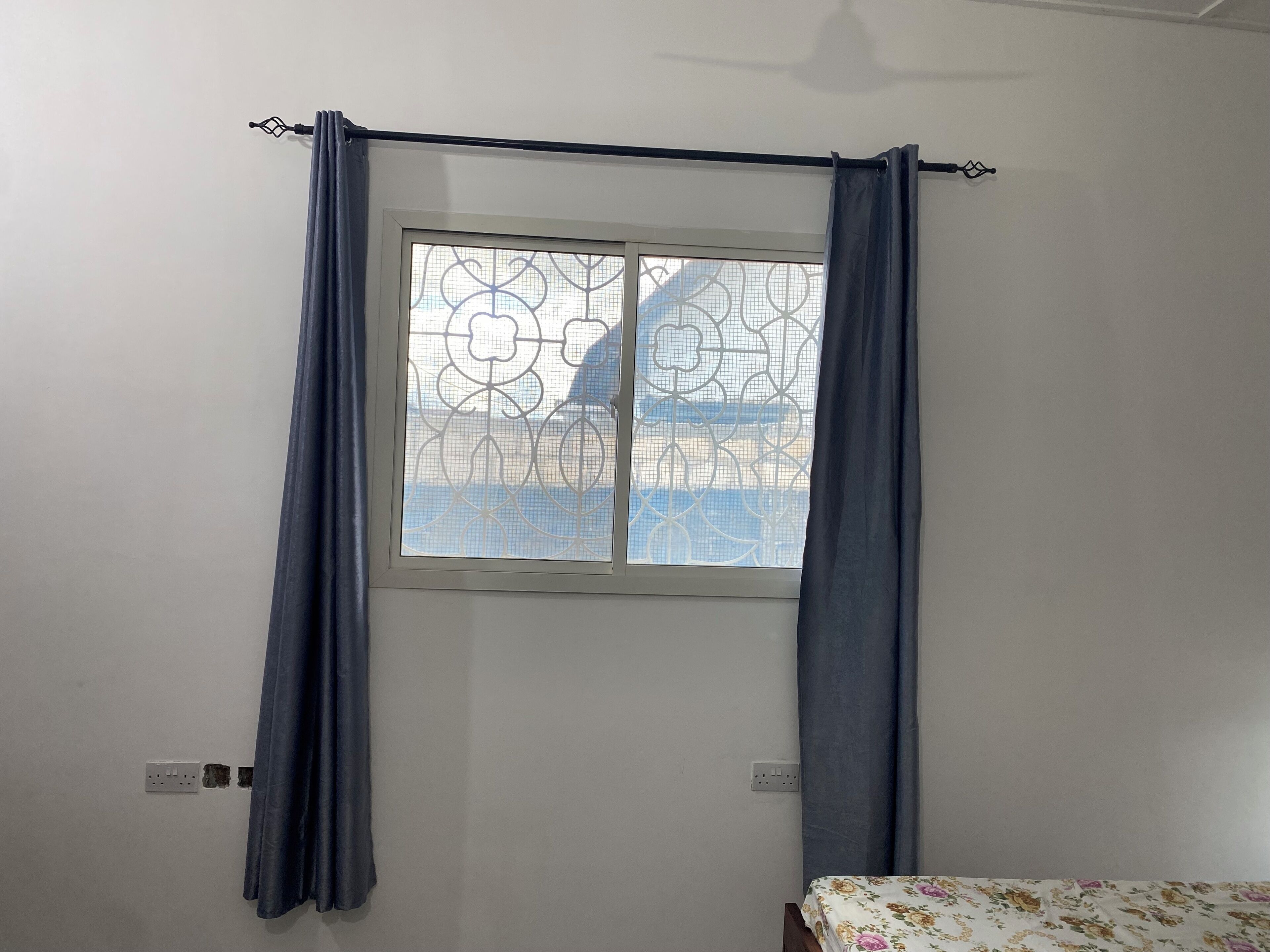 Ein Zimmer mit einem Fenster, das mit einem Metallgitter und blauen Vorhängen verziert ist, einem Bett mit einer blumigen Tagesdecke und einer Wandsteckdose.
