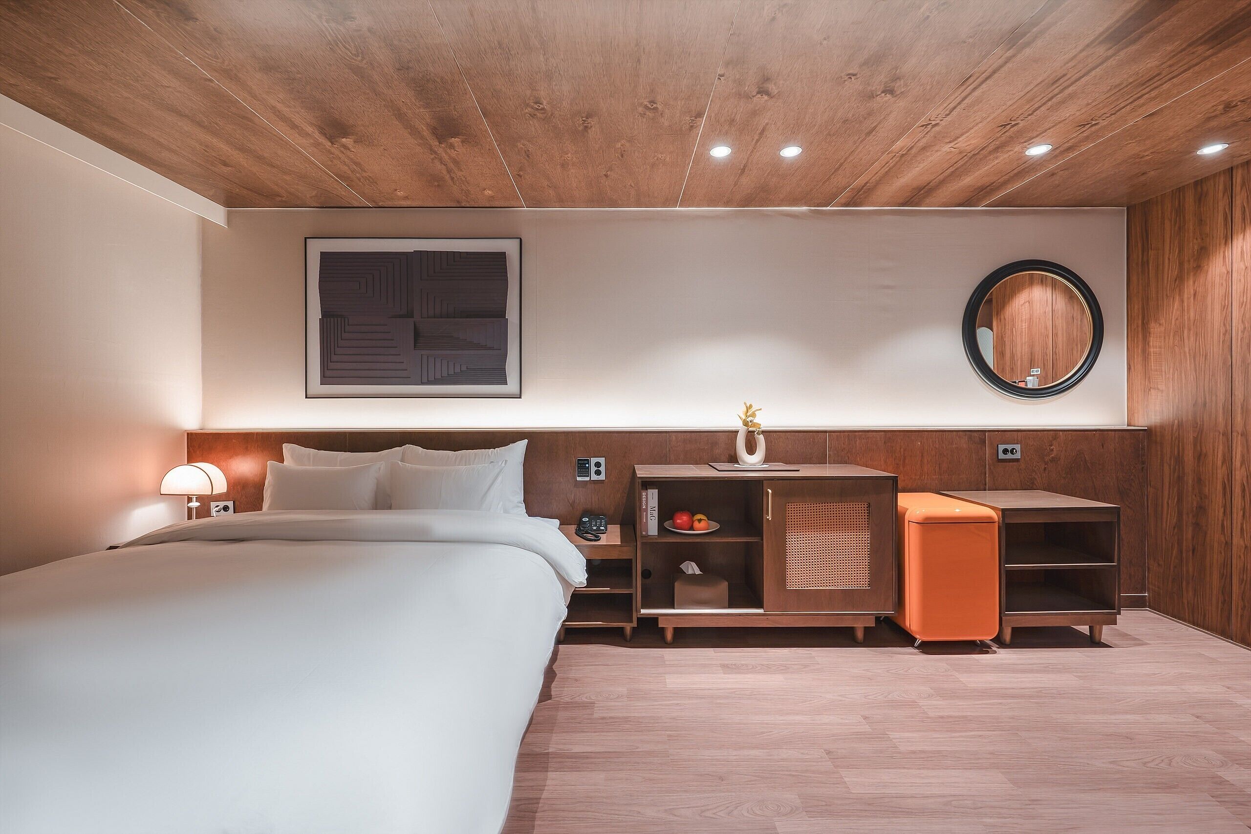 A modern hotel room with a wooden ceiling, a large bed, a nightstand, a dresser, a mirror, and a small orange cabinet.