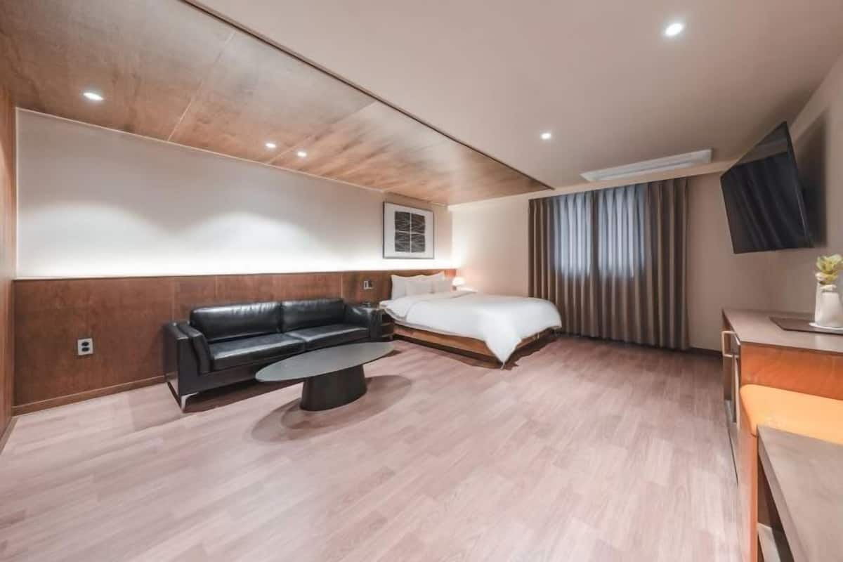 Moderni hotellihuone, jossa on puukoristeinen seinä, musta nahkasohva, valkoisilla tekstiileillä varustettu sänky, pieni pyöreä sohvapöytä ja seinälle asennettu litteänäyttöinen televisio.