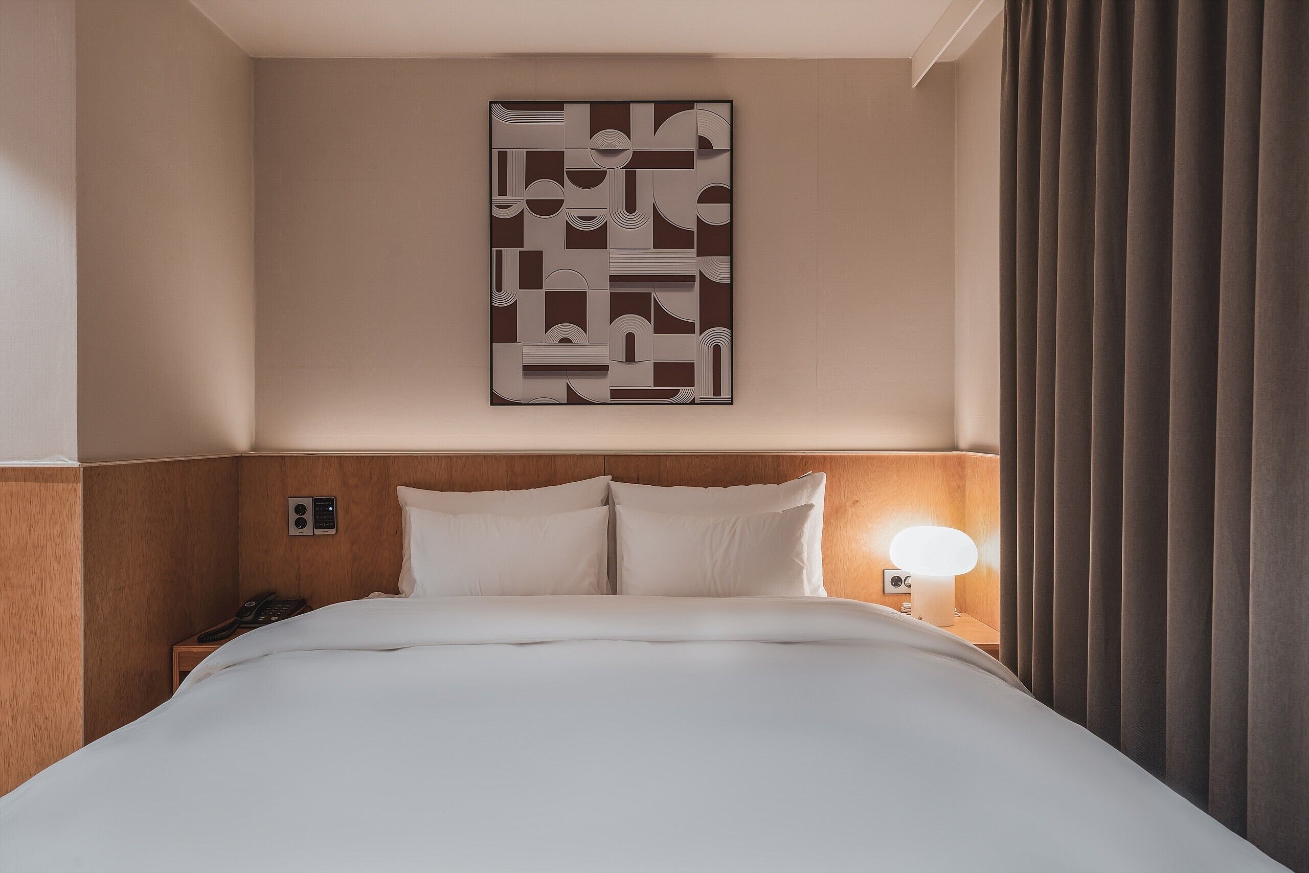 A hotel room with a large abstract artwork on the wall, a neatly made bed with white linens, a bedside table with a lamp, and a telephone.