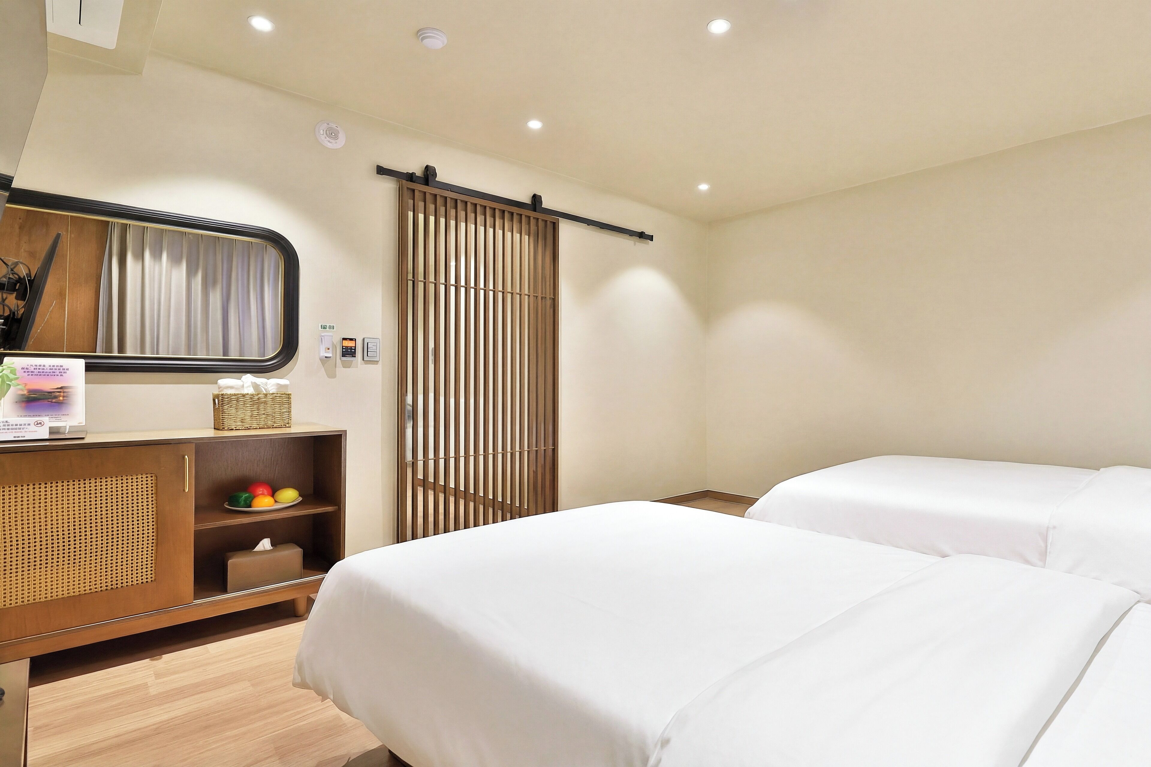 Ein Hotelzimmer mit einem Bett, einem Nachttisch mit Spiegel, einer kleinen Ablagefläche mit Früchten und einer Schiebetür.