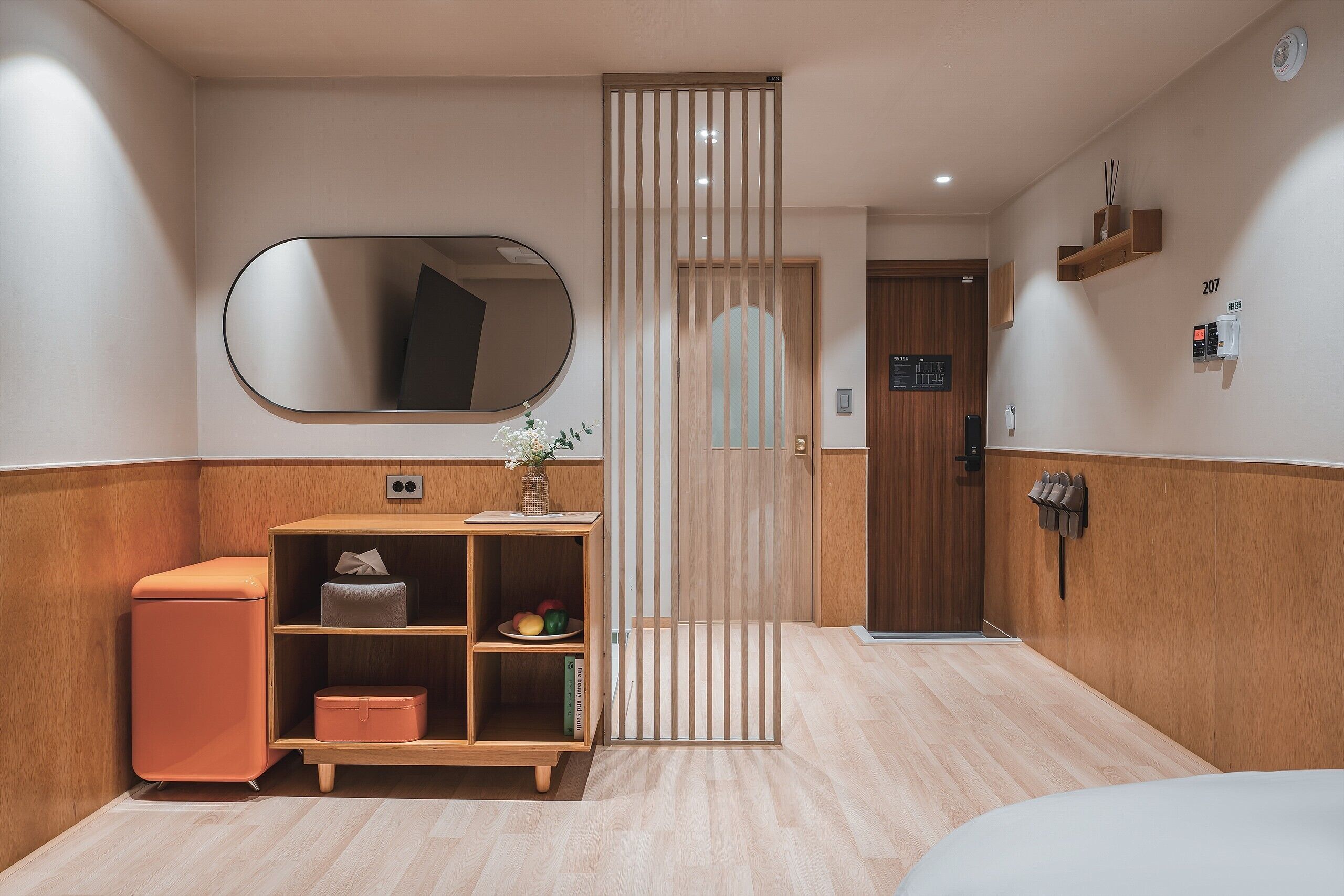 A hotel room with a wooden cabinet, a mirror, a small table with a vase, and a door with a number sign.