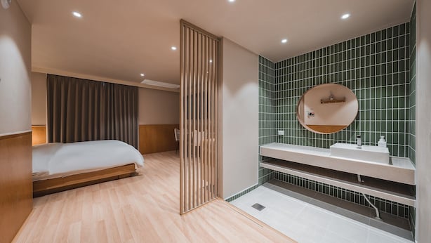 Una habitación de hotel moderna con una cama, un separador de madera, un baño con lavamanos y espejo, y paredes con azulejos verdes.