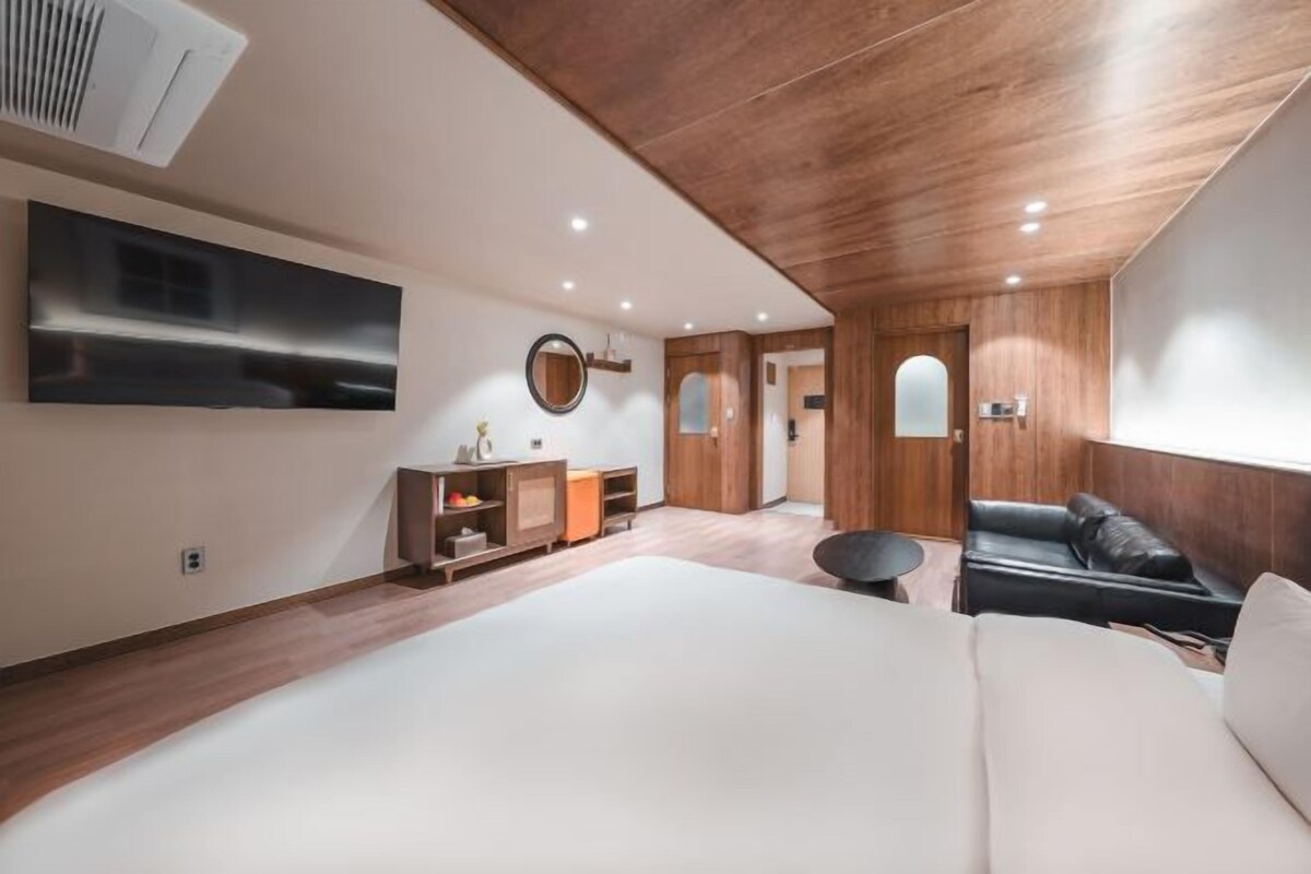 Una habitación de hotel moderna con una cama grande, techo de madera, televisor de pantalla plana, una mesita redonda pequeña y un sofá de cuero negro.