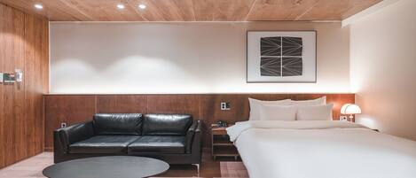 A modern hotel room with a wooden ceiling, a black leather sofa, a round coffee table, a bed with white linens, and a wall-mounted artwork.