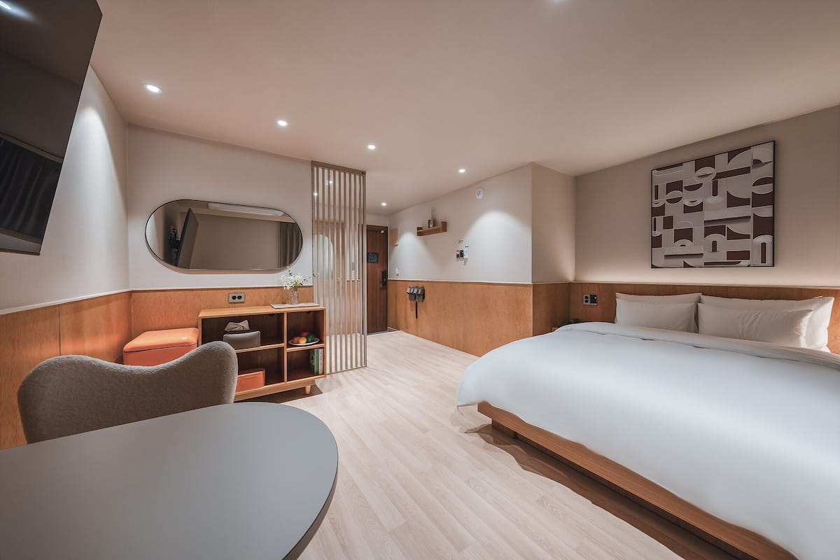 Moderni hotellihuone, jossa on suuri sänky, työpöytä ja tuoli. Huoneessa on myös puinen kaappi, jossa on peili ja hyllyt.