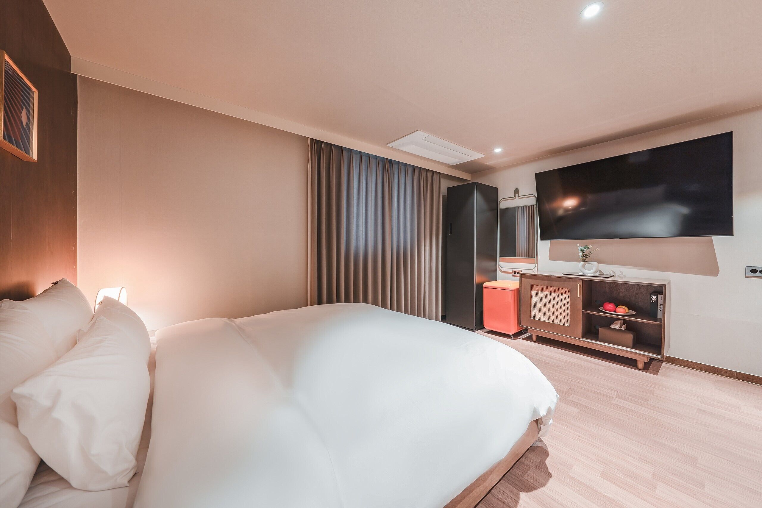 A modern hotel room with a large bed, a flat-screen TV mounted on the wall, a wooden nightstand, and a large window with curtains.