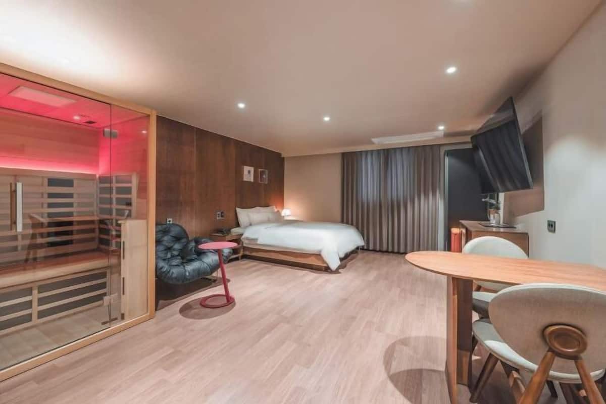 Moderni hotellihuone, jossa on sänky, työpöytä, tuoli, televisio ja lasiseinä.