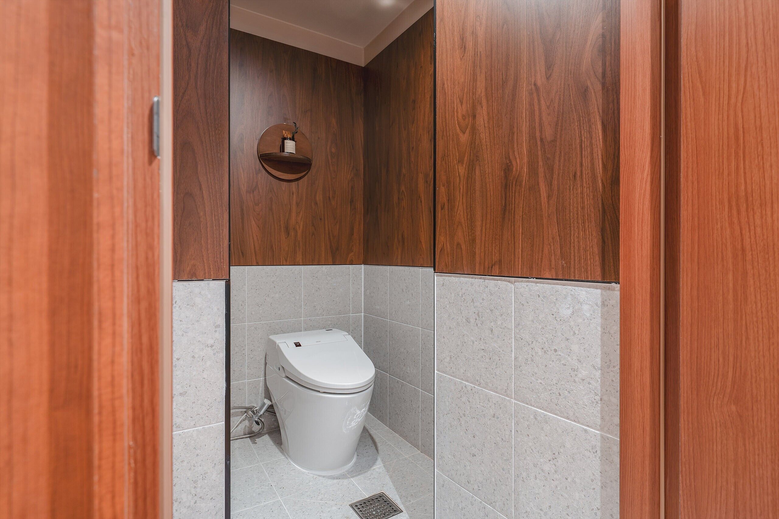 Een kleine, afgesloten badkamer met een toilet, een houten wastafelmeubel en een wandplank.