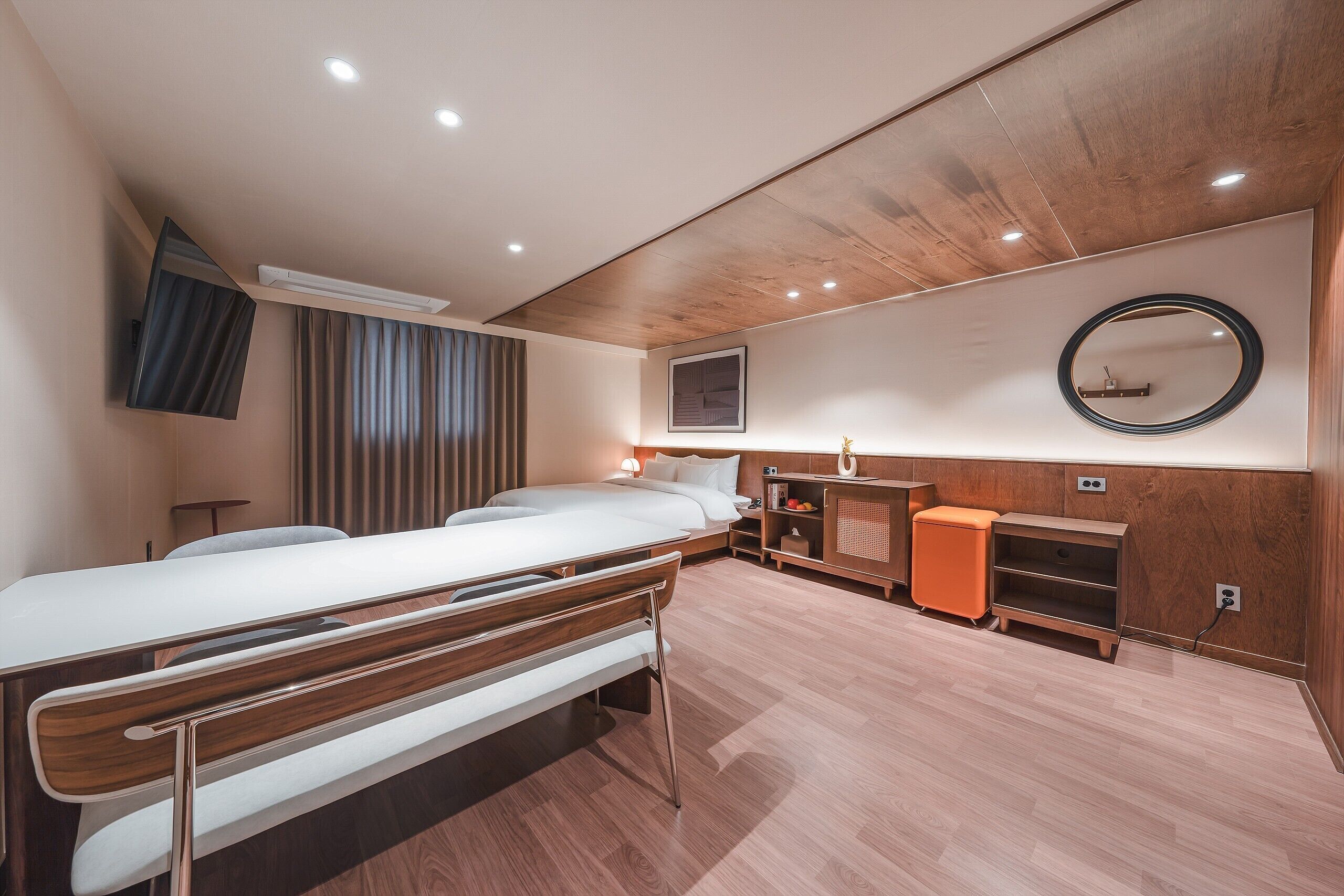 A modern hotel room with a wooden ceiling, a bed, a desk, a chair, and a small orange ottoman.