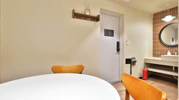 A hotel room with a round table, two chairs, a door, a mirror, a sink, and a fire extinguisher.