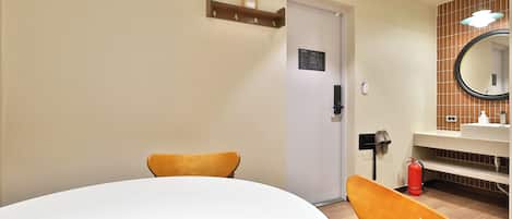A hotel room with a round table, two chairs, a door, a mirror, a sink, and a fire extinguisher.