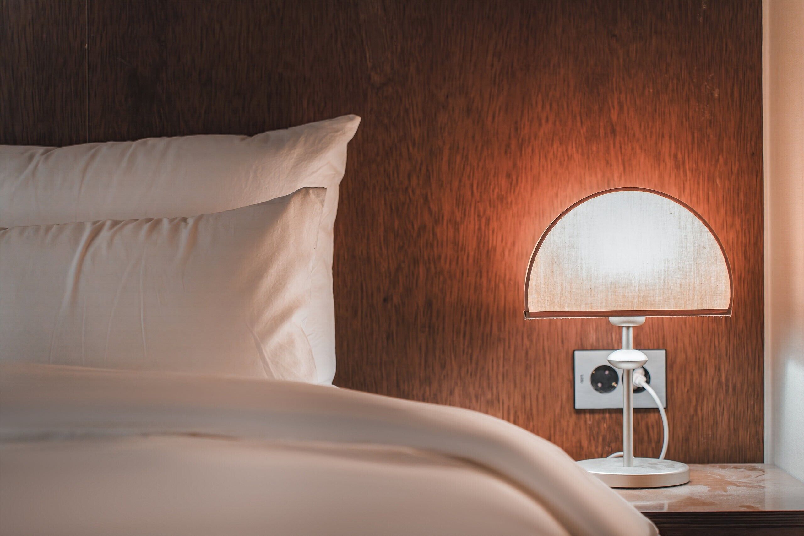 A neatly made bed with a white pillow and a white blanket, a white table lamp with a round shade, and a wooden wall in the background.