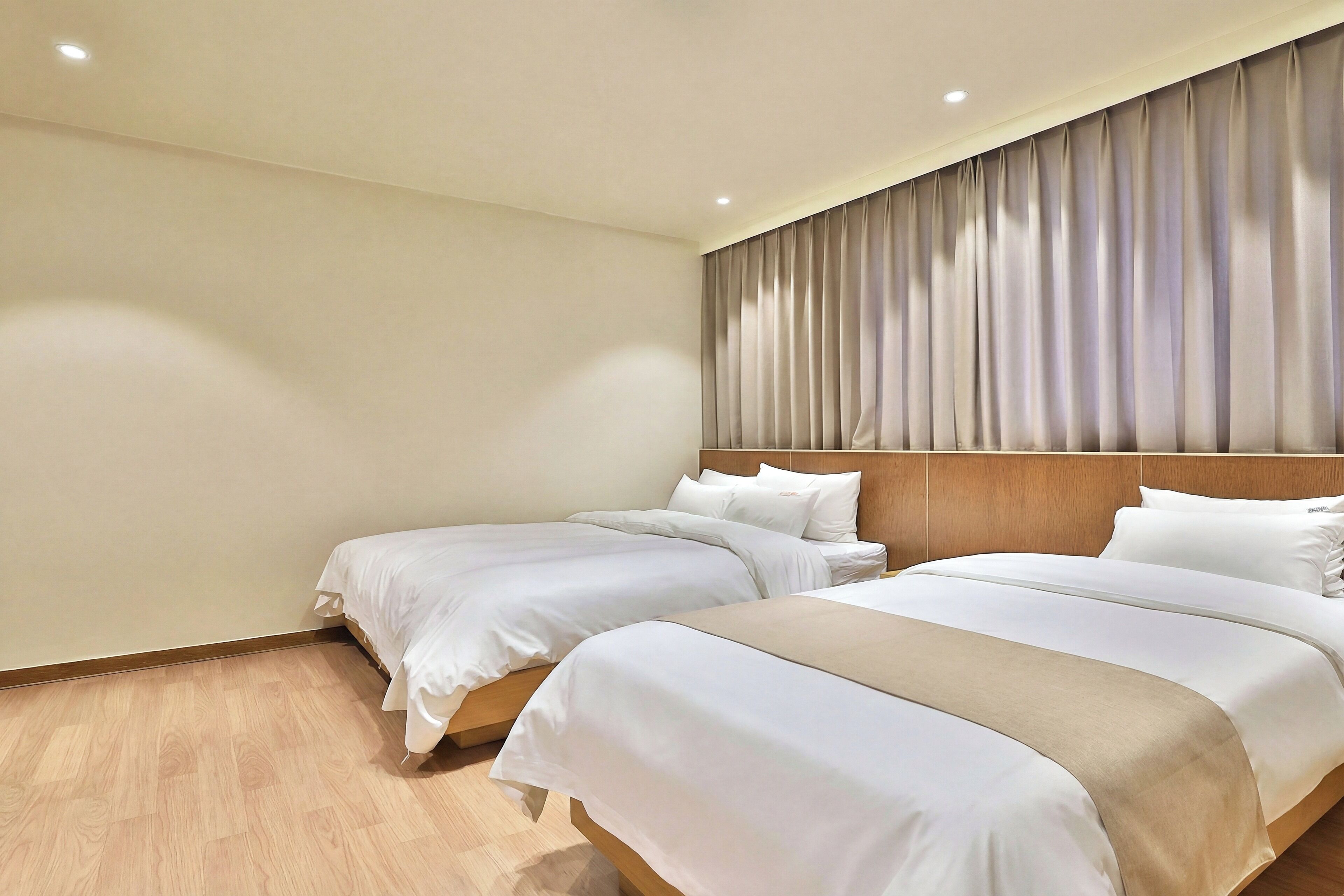 Ein Hotelzimmer mit zwei Betten, Holzboden, einem Vorhangverhang an der Kopfteil und eingelassener Deckenbeleuchtung.