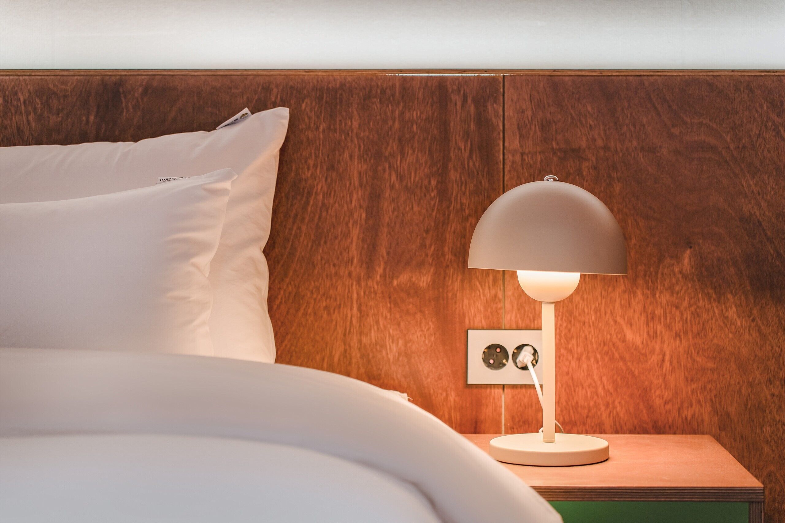 A neatly made bed with white linens and pillows, a bedside table with a lamp, and a wooden headboard.
