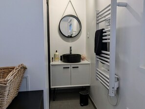 A small bathroom with a round mirror, a sink, a white cabinet, a black trash bin, and a white towel rack.