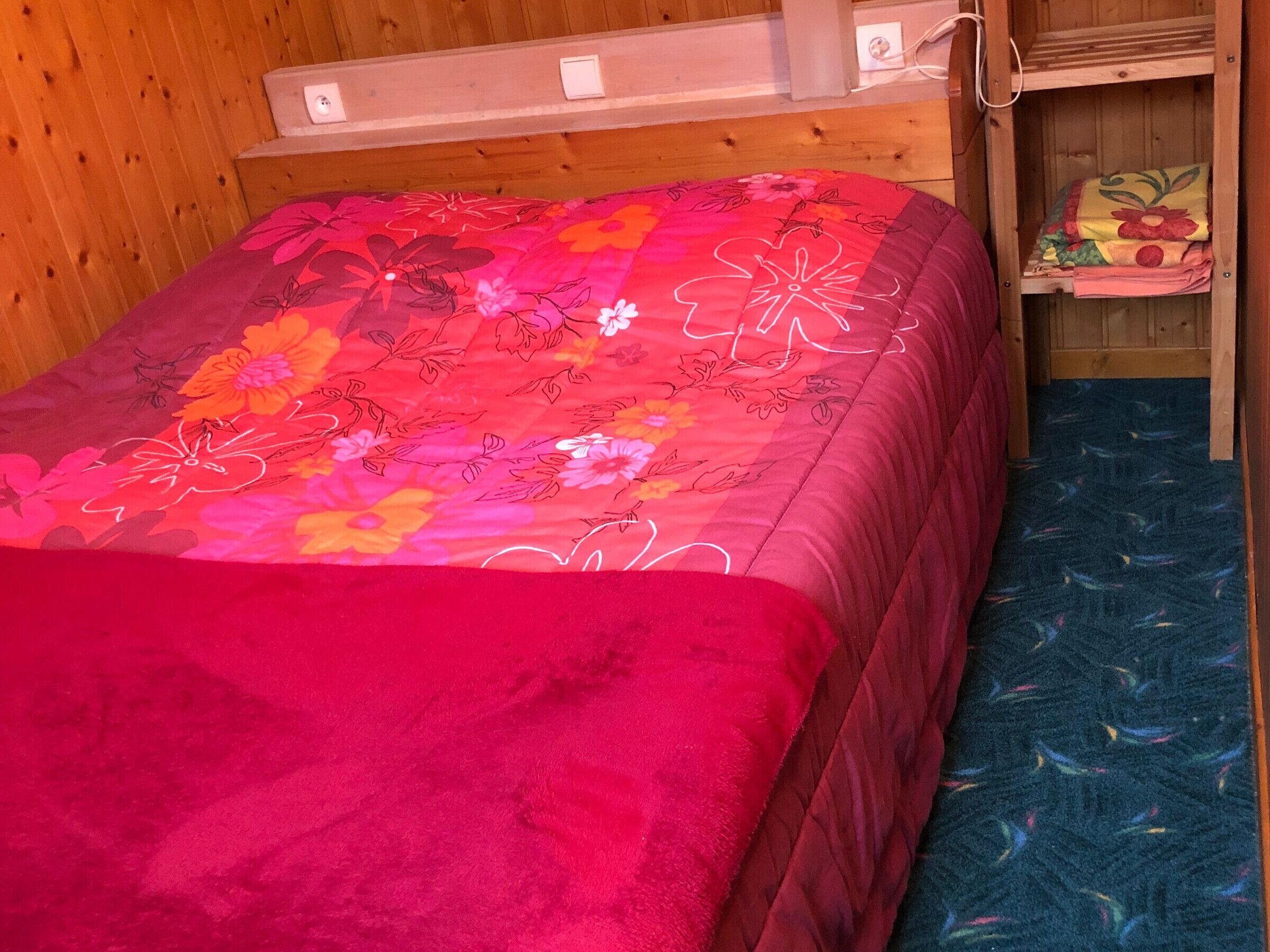 A single bed with a floral bedspread and a red blanket, a wooden bunk bed with a blue patterned mattress, and a wooden wall with a power outlet.