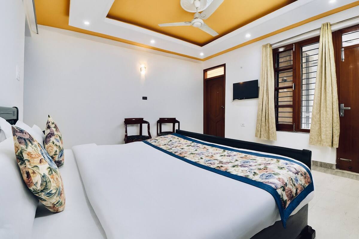 Un dormitorio con cama, dos sillas, ventilador de techo, ventana con cortinas y televisión en la pared.