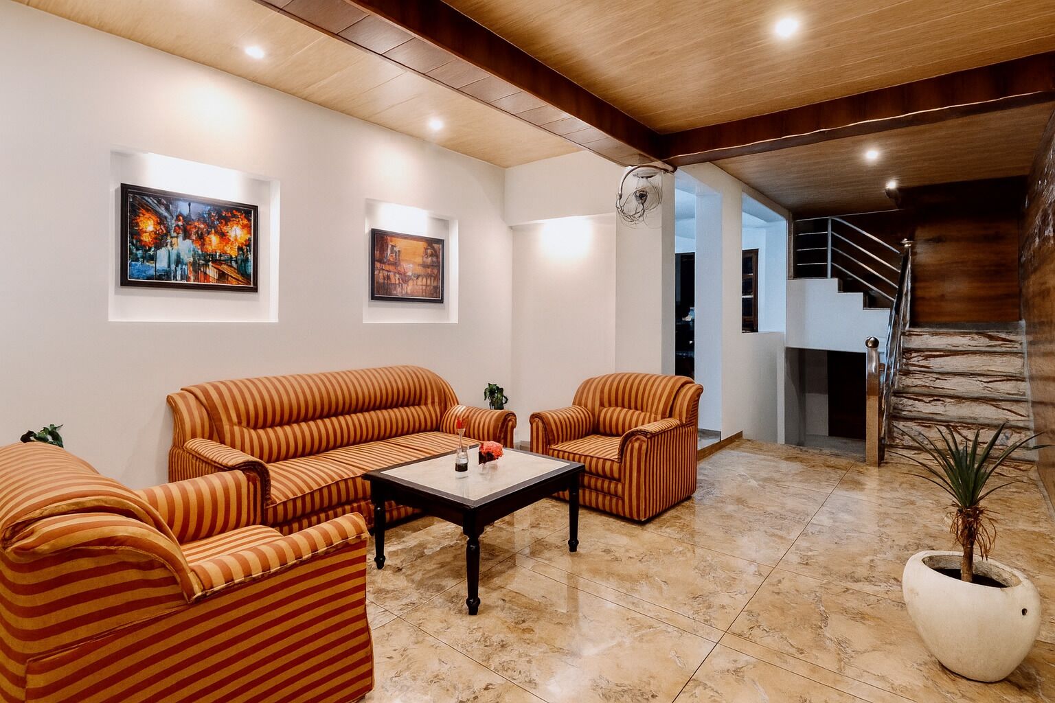 A living room with striped sofas, a coffee table, and a potted plant. There are framed artworks on the walls and a staircase in the background.