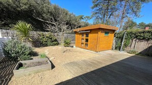 A wooden cabin with a small deck, surrounded by a garden with various plants and a sandy area. - 3-bedroom house near the beach with private garden and parking (Longeville-sur-Mer)