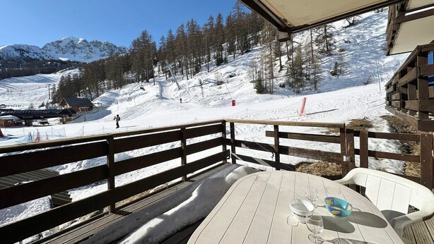 A wooden deck with a table and chairs, overlooking a snowy mountain landscape with ski lifts and a lodge.