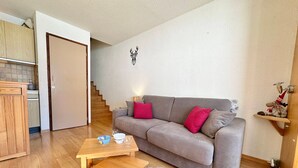 A living room with a grey sofa, a wooden coffee table, and a small rug. A kitchen area with a wooden cabinet and a tiled backsplash is visible in the background.