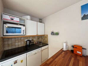Une cuisine compacte équipée d’un four à micro-ondes, d’une machine à café et d’un lave-vaisselle. On y trouve une petite étagère avec les ustensiles de cuisine essentiels et une poubelle.
