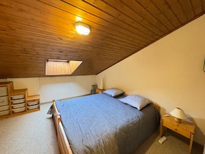 A bedroom with a sloped ceiling, a skylight, a bed with a patterned bedspread, a bedside table with a lamp, and a nightstand with a lamp.