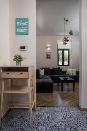 A living room with a sofa, a coffee table, a desk, a potted plant, a wall-mounted thermostat, a framed picture, and a window with a view.