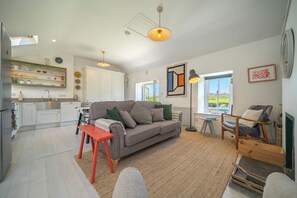 Een moderne keuken met witte kasten, een zithoek met een grijze bank, en uitzicht op de omgeving door een raam.