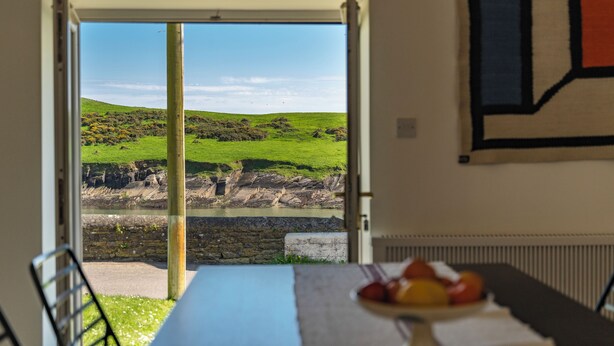 A dining area with a table set for two, a view of a green landscape through an open door, and a wall-mounted painting.