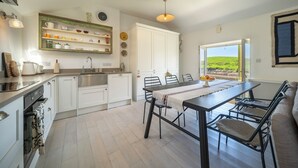 A modern kitchen with white cabinets, a dining table with chairs, and a view of a green landscape through a large window.