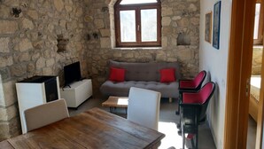 Una habitación con paredes de piedra, una mesa de madera, sillas blancas, un sofá gris con cojines rojos y un televisor sobre un mueble.