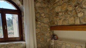 Un dormitorio con pared de piedra, cabecera de madera, ventana con cortinas y cama con colcha blanca.