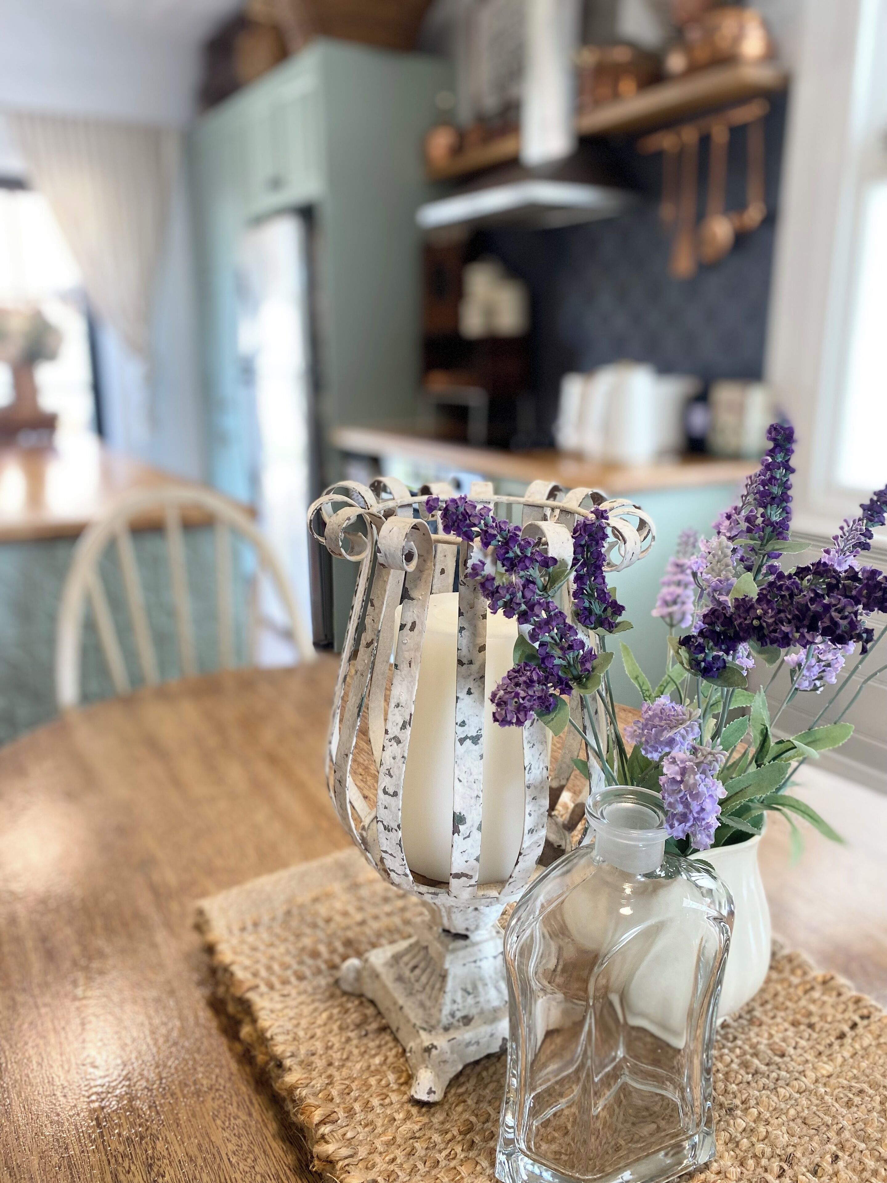 A rustic wooden table with a white vase containing purple flowers and a clear glass bottle.