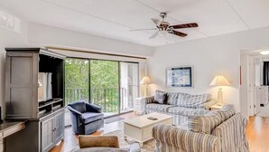 Un salon spacieux avec une grande fenêtre, un ventilateur de plafond et un téléviseur posé sur un meuble.