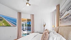 Un dormitorio con cama, ventilador de techo, un cuadro, ventana con cortinas y vistas a una piscina y playa.