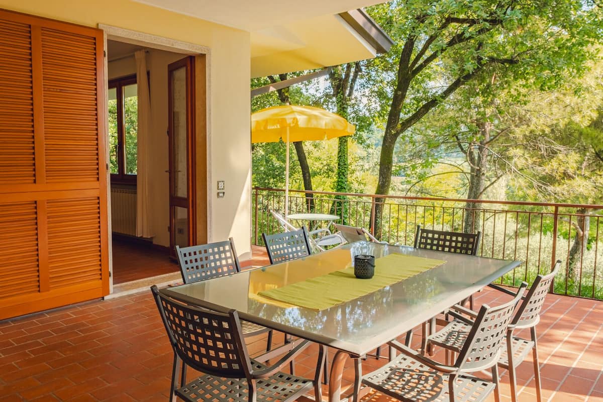 Une terrasse avec une table et des chaises en verre, un parasol jaune et une vue sur les arbres.