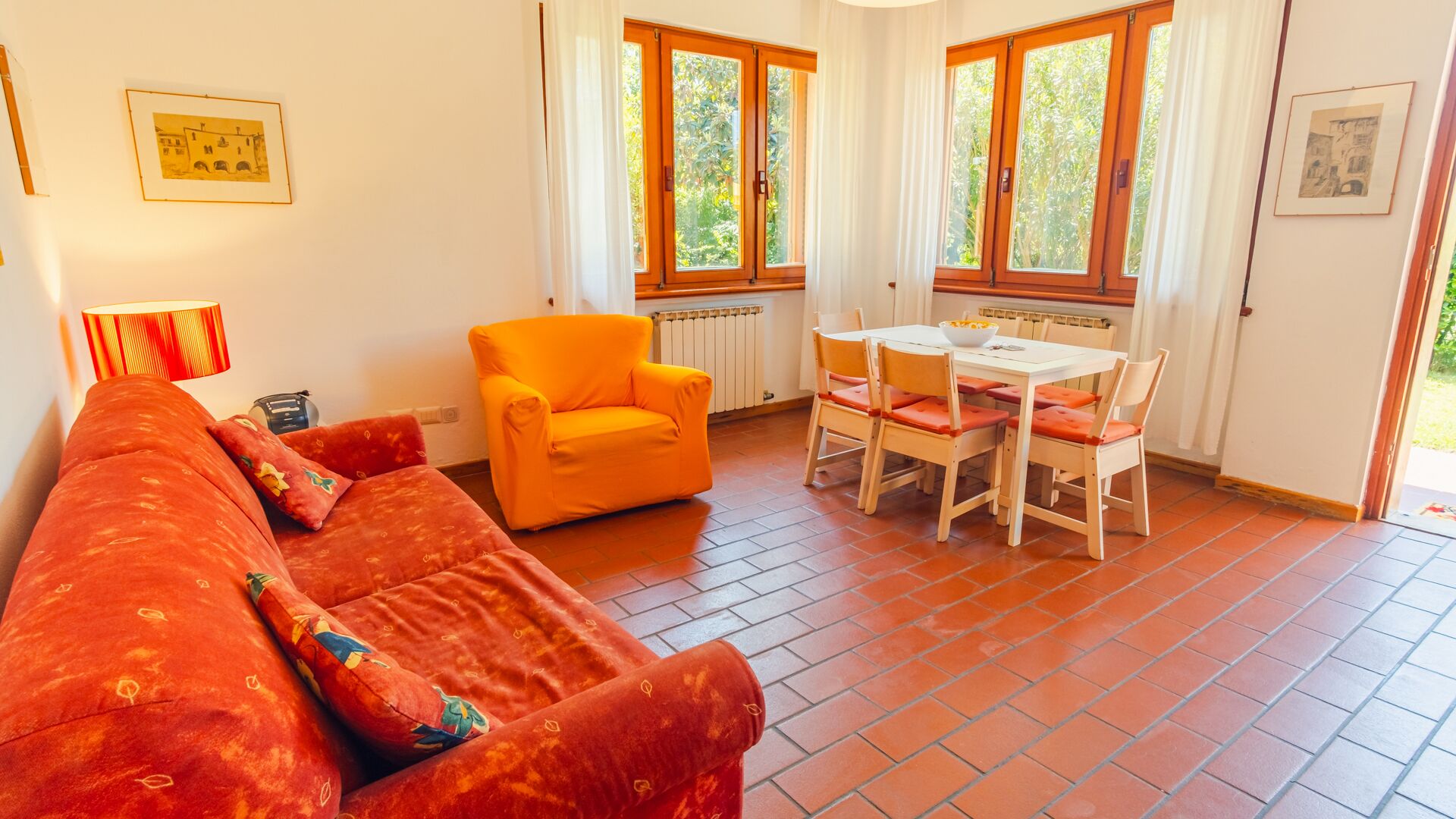 Ein Wohnzimmer mit einem roten Sofa, einem orangefarbenen Sessel, einem Esstisch mit Stühlen und großen Fenstern mit Vorhängen.