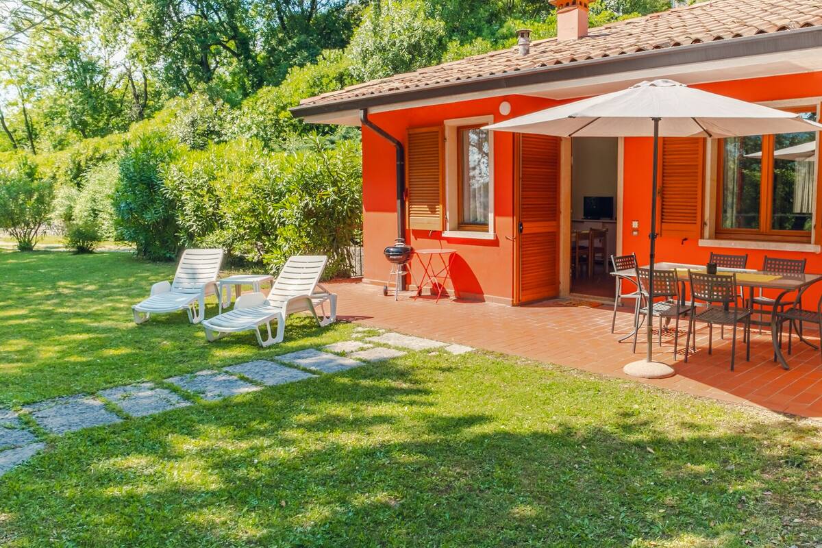 Une maison à l’extérieur rouge, une terrasse avec une table et des chaises, et une pelouse avec des chaises longues.